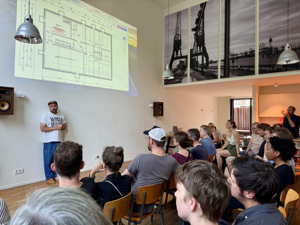 Baugruppentreffen im Juli 2025: Ein Mitglied stellt vor vollem Saal einen Grundriss vor